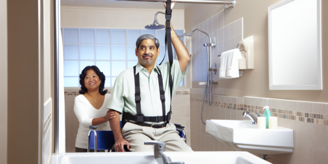 A person in a wheelchair being lifted by an overhead ceiling lift with a caregiver standing behind in a bathroom.